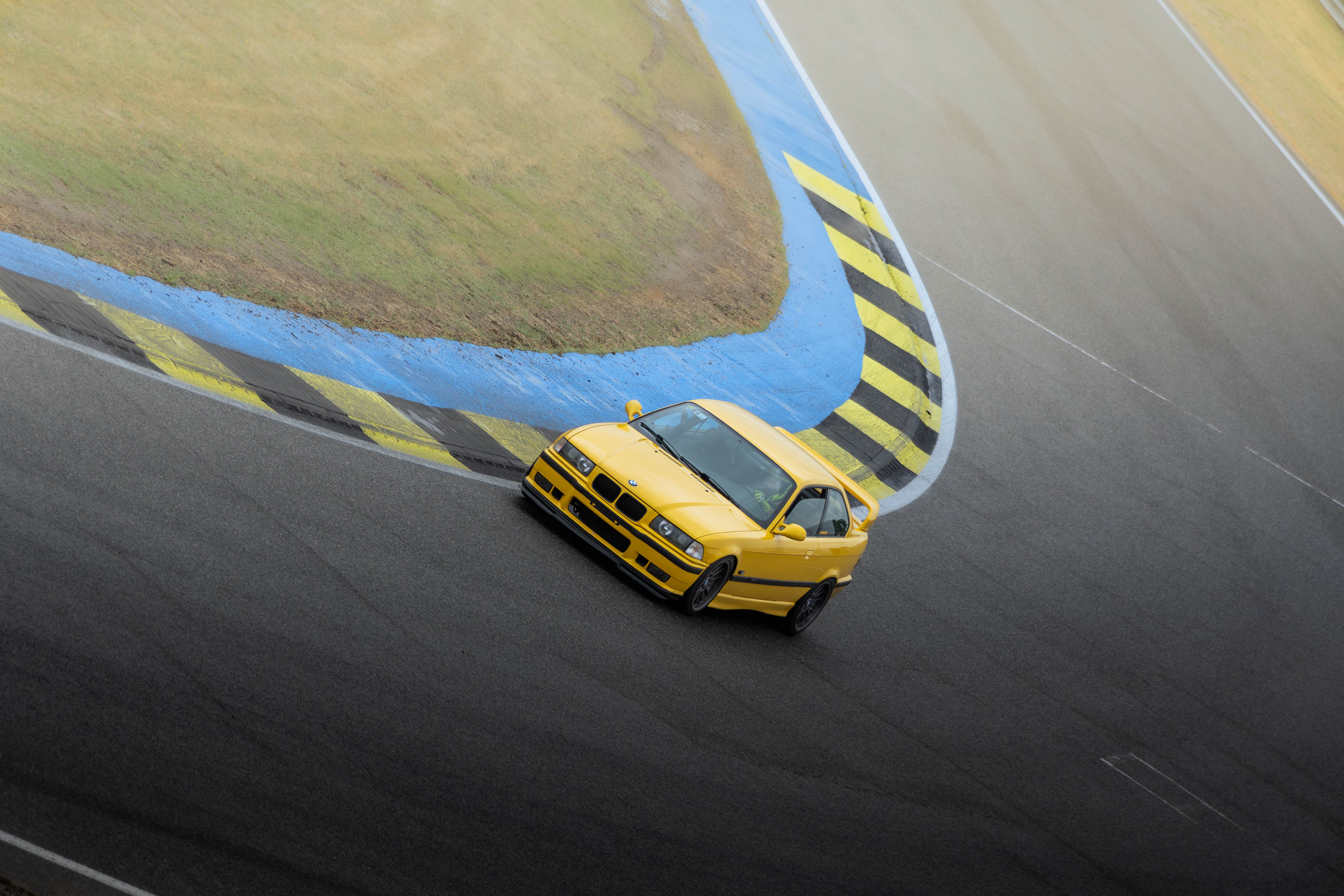 BMW E36 M3 amarillo en una curva azul y amarilla de Le Mans, tomado desde arriba.